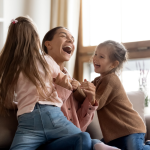 Mother laughing and bonding with two young daughters at home during playful family time – representing positive family moments supported by ABA therapy in Greenville, SC.